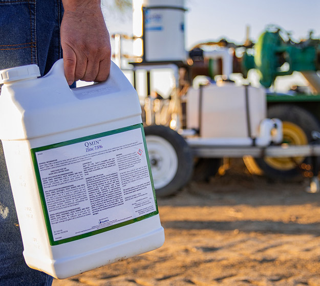 Farmer holding a bottle of Qmin Zinc Fertilizer with a Tank Sprayer in the Background
